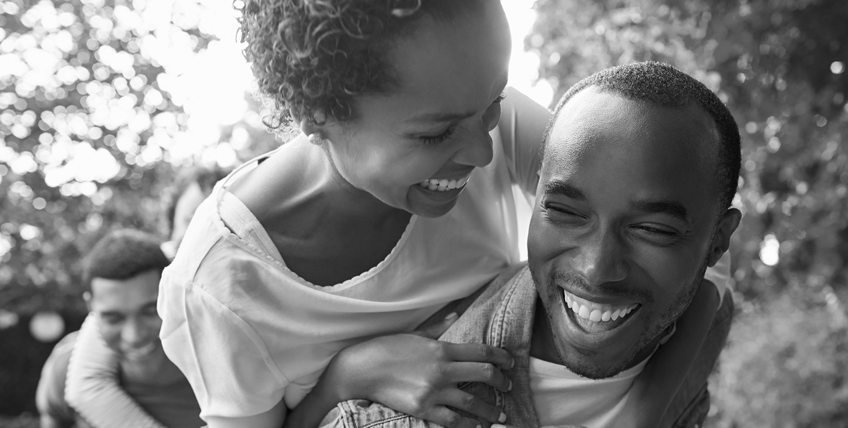 Smiling woman leaning on the back of a smiling man in an outdoor setting with trees in the background